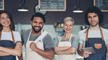 Four smiling people with aprons stand in front of a cafe counter.